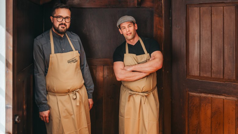 2 Personen mit Schürze stehen vor einer mit Holz vertäfelten Wand.