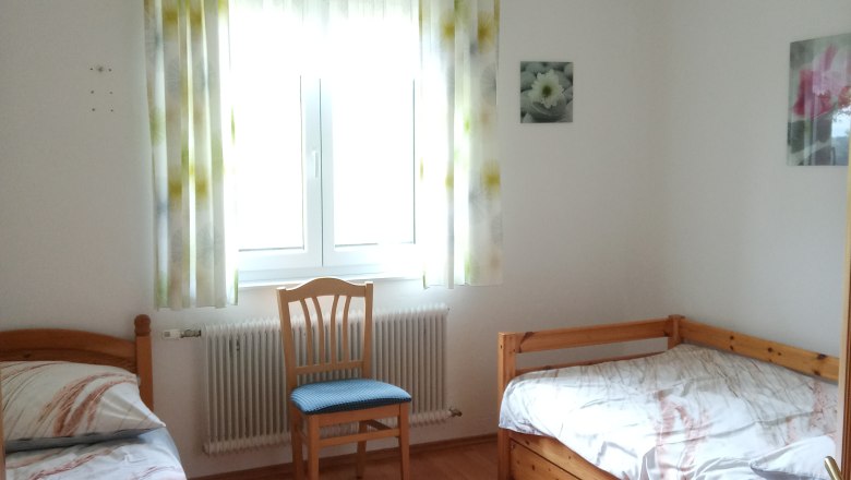Ein einfach eingerichtetes Schlafzimmer mit zwei Einzelbetten, einem Stuhl und einem Fenster mit Vorhängen.