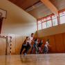 Fünf Jugendliche rennen in einer Turnhalle mit Basketballkorb und Handballtor.