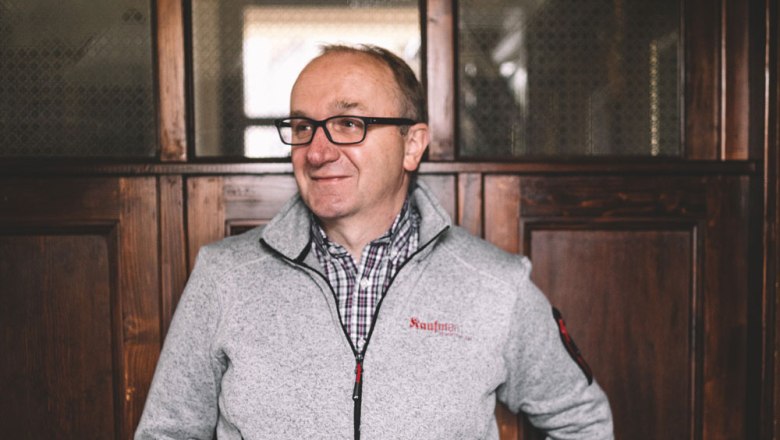 A man with glasses and a gray jacket stands in front of a wooden wall.