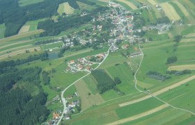 Luftaufnahme einer ländlichen Ortschaft mit Feldern und Wäldern.