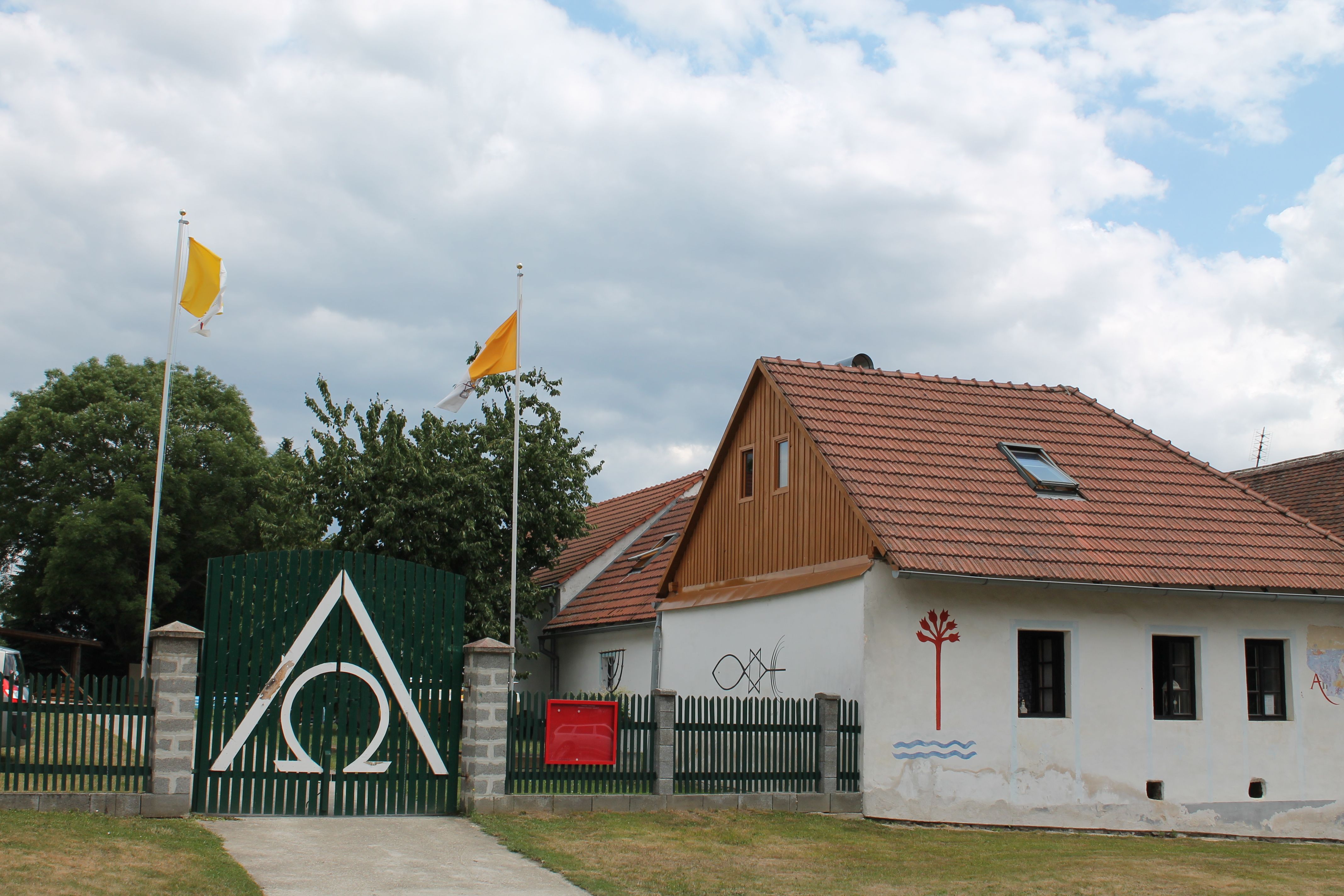 Ein Haus mit roten Dachziegeln, grüner Zaun mit Alpha-Omega-Symbol, zwei gelbe Fahnen und Wandmalereien.