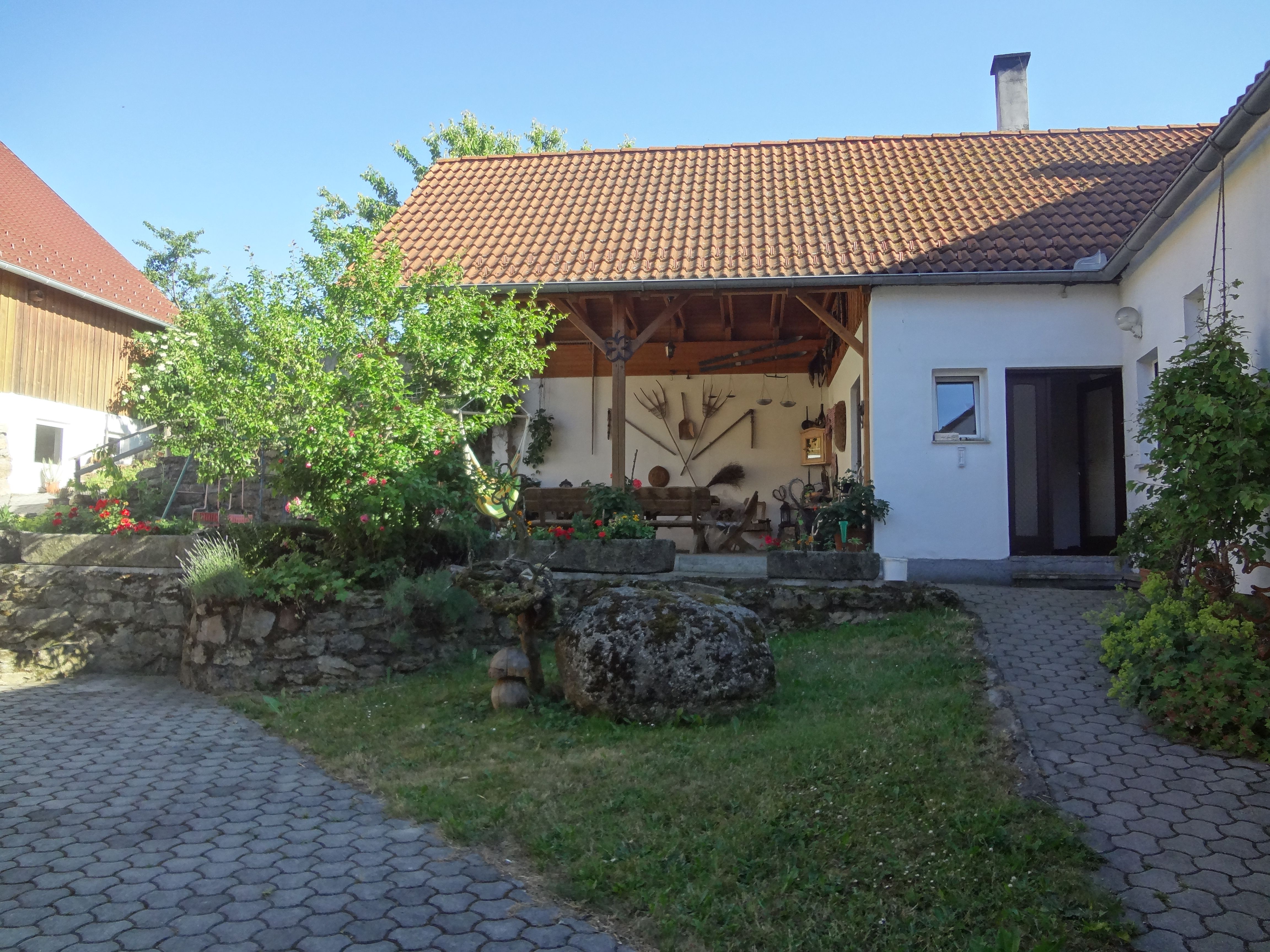 Hof mit weißem Haus, rotem Ziegeldach, überdachter Terrasse, Garten und gepflasterten Wegen bei sonnigem Wetter.