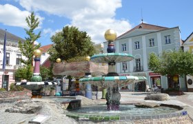 Ein bunter Brunnen im Hundertwasser-Stil mit goldenen Kugeln und fließendem Wasser, umgeben von Gebäuden und Bäumen.