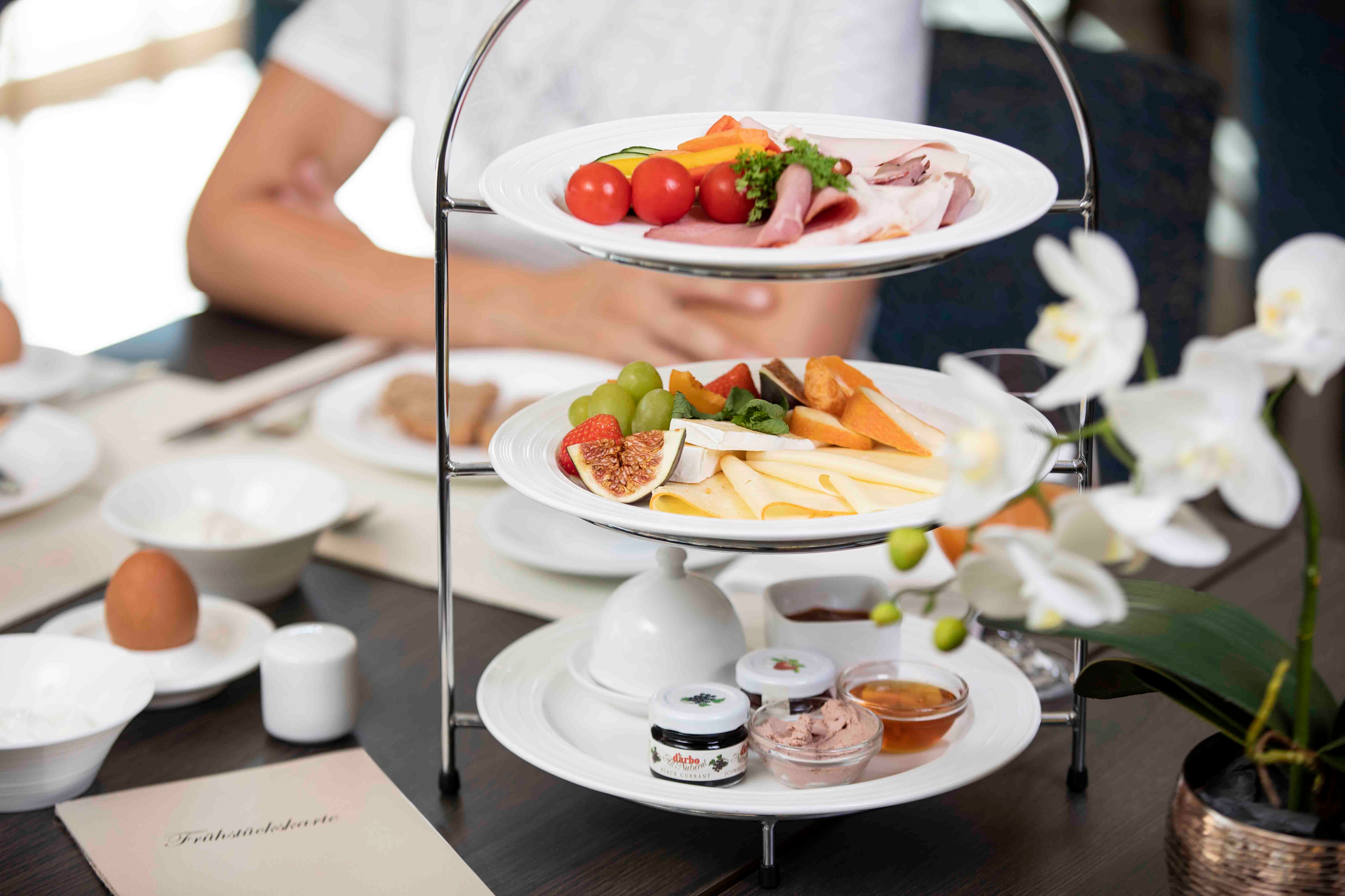 Ein Frühstückstisch mit einer Etagere voller Käse, Aufschnitt, Obst und Brotaufstrichen. Im Vordergrund eine weiße Orchidee.