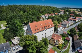 Luftaufnahme eines großen Gebäudes mit rotem Dach, umgeben von Bäumen und kleineren Häusern in einer ländlichen Gegend.