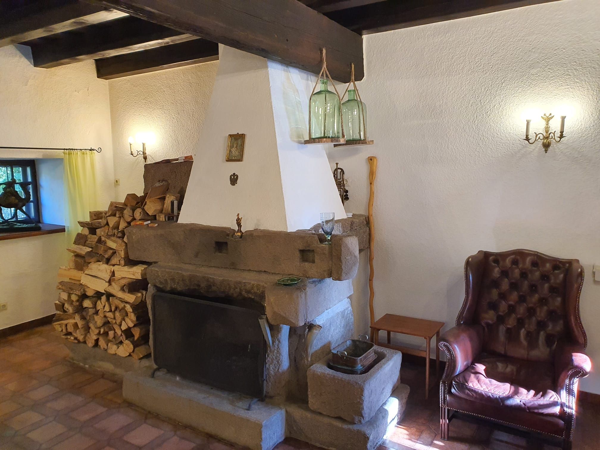 Ein rustikales Wohnzimmer mit einem großen Kamin, einem Stapel Holz, einem Ledersessel und zwei Wandlampen.
