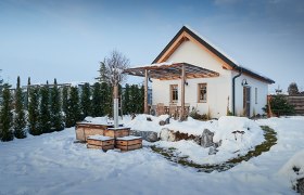 Ein Chalet im Schnee mit Terrasse und Gartenmöbeln, umgeben von Bäumen.