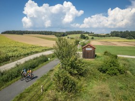 Die sanften H&uuml;gel und weitl&auml;ufigen Felder laden zu einer erfrischenden Radtour ein. Radfahrer genie&szlig;en die idyllische Landschaft, w&auml;hrend die Sonne &uuml;ber den Horizont strahlt und die Wolken sanft am Himmel treiben. Ein perfekter Tag, um die Natur in ihrer vollen Pracht zu erleben.