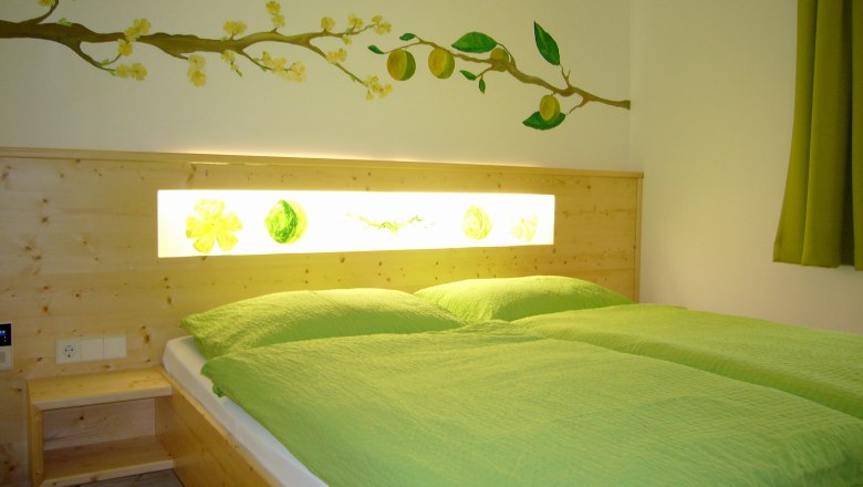 Ein gemütliches Zimmer mit einem Doppelbett, grüner Bettwäsche und einer Wanddekoration mit gelben Blüten und Früchten.