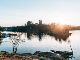 Die sanften Wellen des Stausees reflektieren das goldene Licht der untergehenden Sonne, w&auml;hrend die Silhouette einer alten Burg majest&auml;tisch im Hintergrund thront. Umgeben von &uuml;ppigem Gr&uuml;n und dem sanften Pl&auml;tschern des Wassers, l&auml;dt dieser Ort zu einem unvergesslichen Naturerlebnis ein.