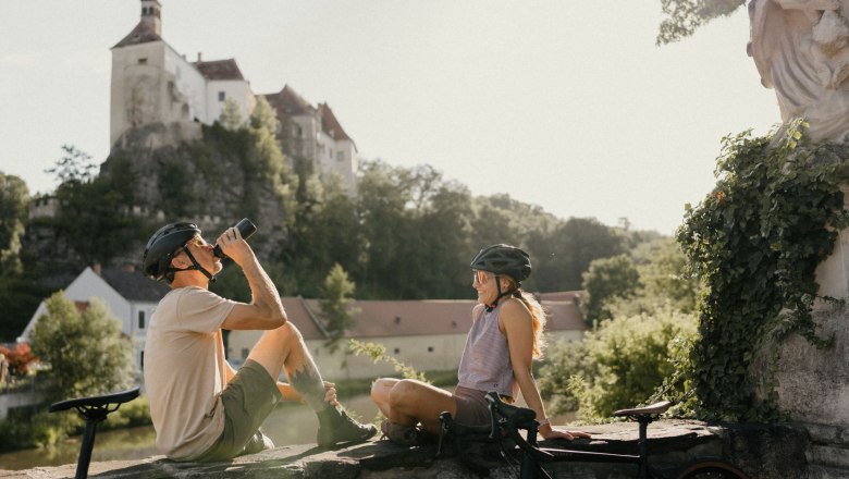 Two cyclists with helmets sit on a wall in front of Raabs Castle.