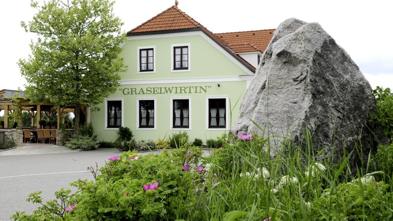 Grünes Gebäude mit der Aufschrift 'Graselwirtin', umgeben von Pflanzen und einem großen Felsen im Vordergrund.