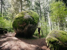 Person steht im Wald vor einem gro&szlig;en, kugelf&ouml;rmigen Felsen, der von Moos bedeckt ist; die sogenannte Weltkugel liegt in der Kraftarena Gro&szlig; Gerungs, umgeben von hohen B&auml;umen.