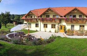 Ein großes gelbes Haus mit roten Dächern und Blumenkästen, umgeben von einem gepflegten Garten mit Sitzbereich und Sonnenschirm.