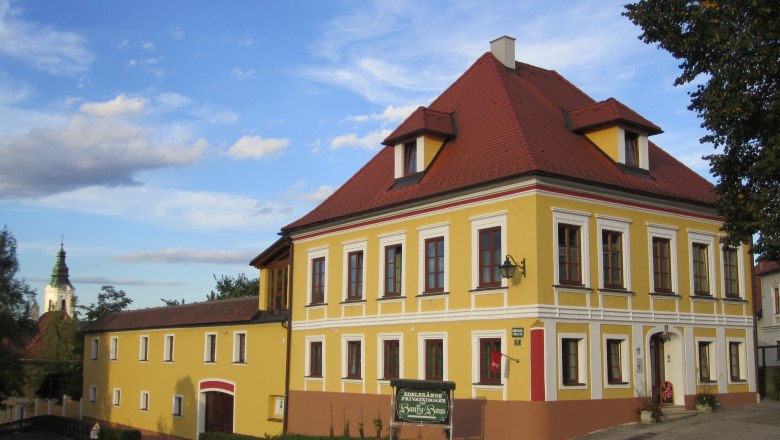Gelbes zweistöckiges Gebäude mit rotem Dach und weißen Fensterrahmen, daneben ein kleineres Gebäude. Im Hintergrund eine Kirchturmspitze.