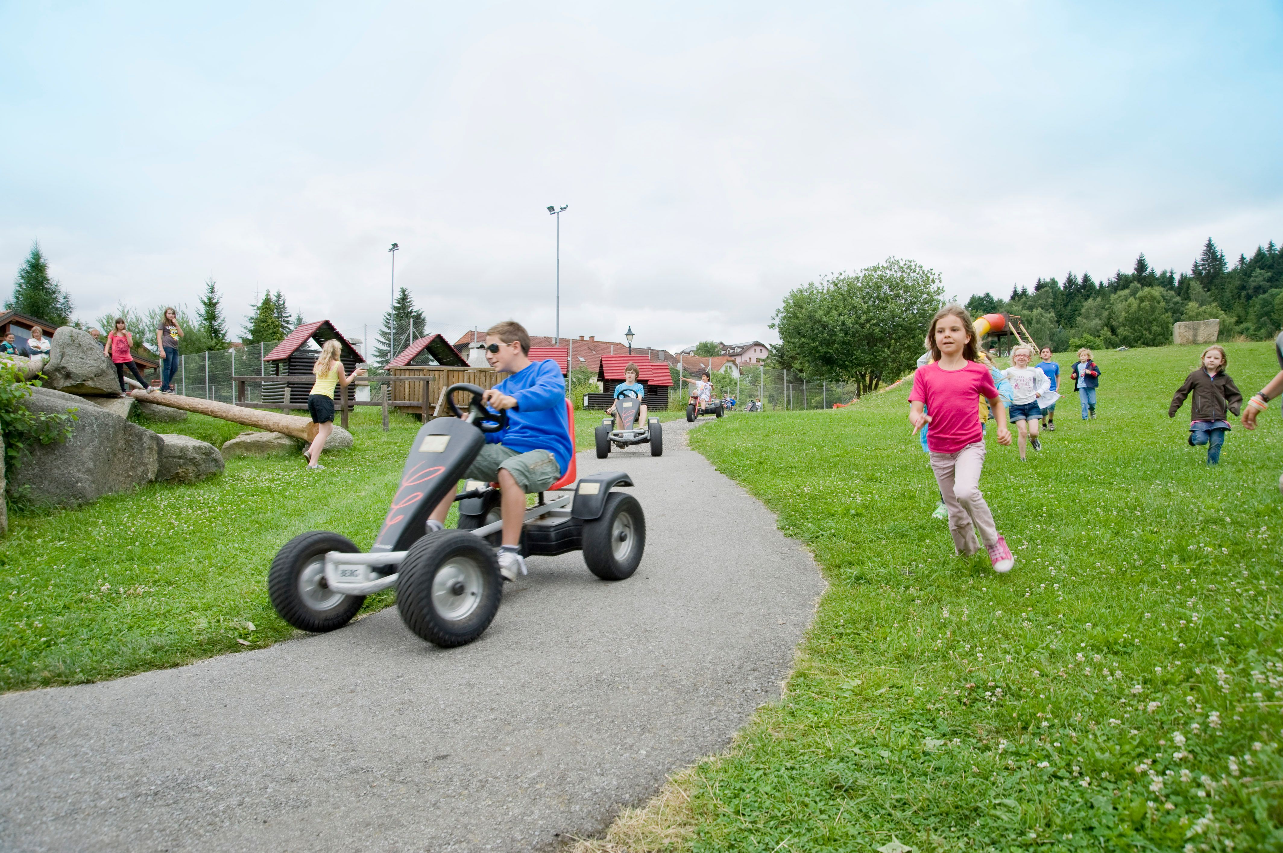 Kinder spielen auf einem Erlebnisspielplatz mit Kettcars und laufen über eine Wiese.