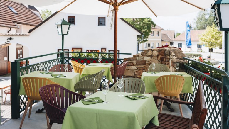 Ein Gastgarten mit grünen Tischdecken und bunten Stühlen unter einem Sonnenschirm, umgeben von Gebäuden und Pflanzen.