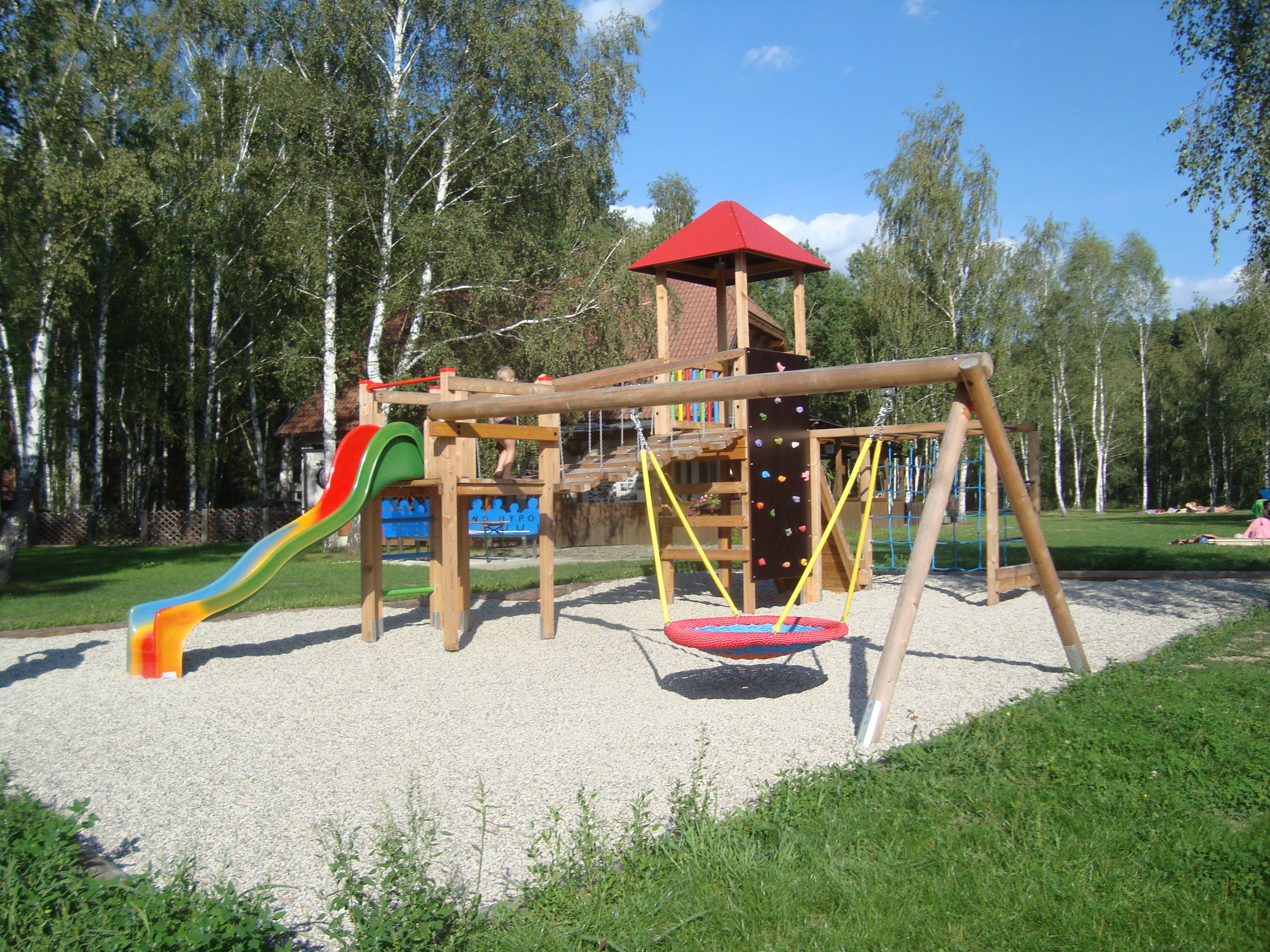 Ein Kinderspielplatz mit Rutsche, Schaukel und Klettergerüst, umgeben von Bäumen.