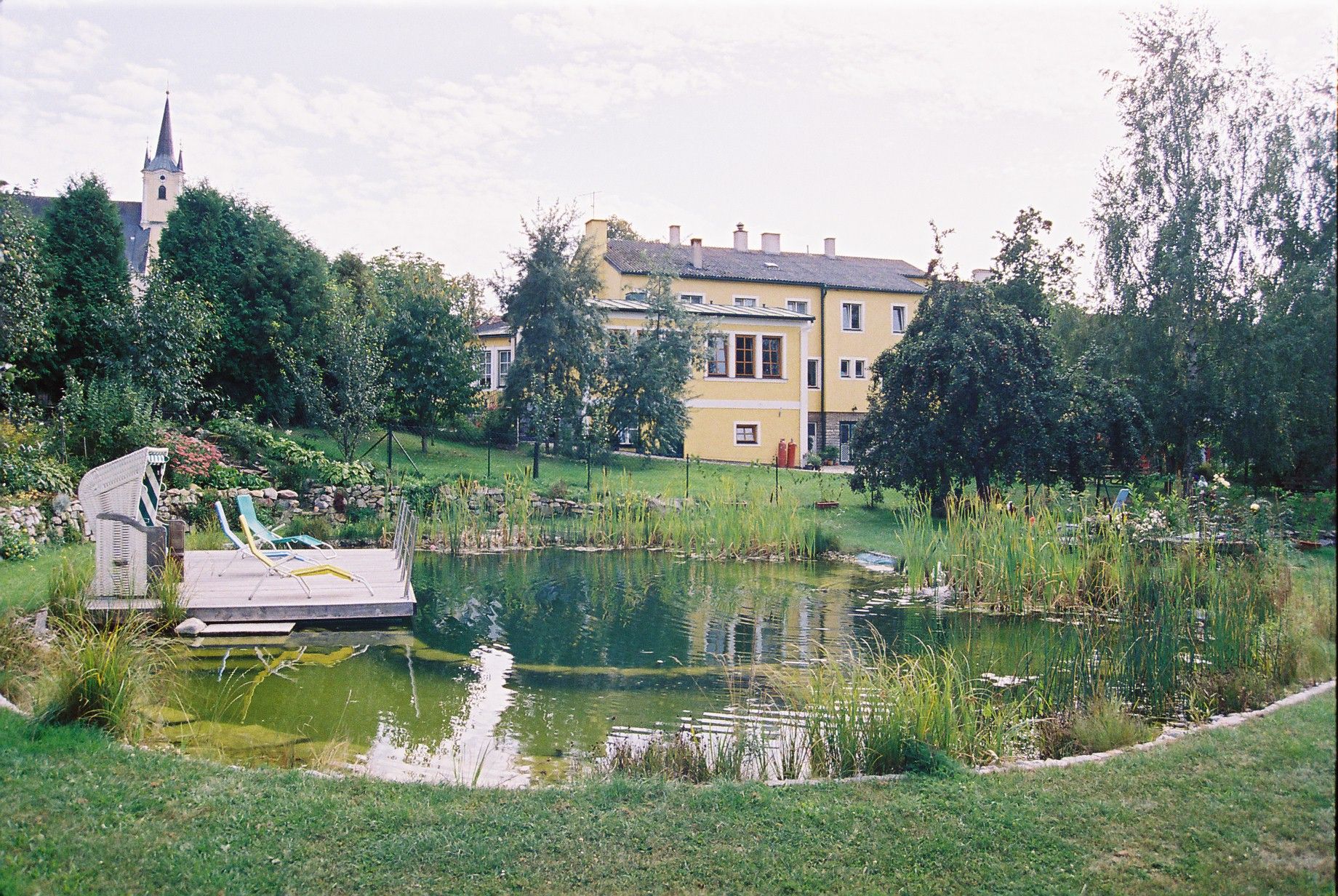 Ein Schwimmbiotop in einem großen Garten mit einem gelben Gebäude im Hintergrund.