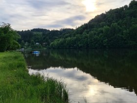 Ein ruhiger See mit bewaldeten H&uuml;geln im Hintergrund und einer leicht bew&ouml;lkten Abendsonne.