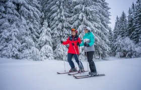 Zwei Skifahrer stehen auf einer verschneiten Piste vor schneebedeckten B&auml;umen.