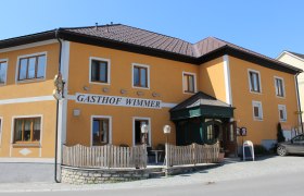 Gelbes Gebäude mit der Aufschrift 'Gasthof Wimmer', Holzzaun und Eingangstür.