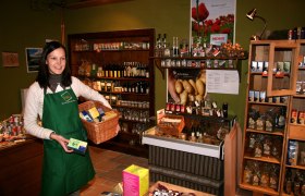 Eine Frau in einem Laden mit grüner Schürze hält einen Korb mit Produkten.