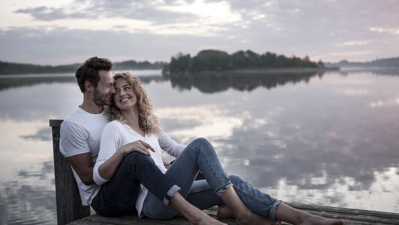 Ein Paar sitzt lächelnd auf einem Steg am See bei Sonnenuntergang.