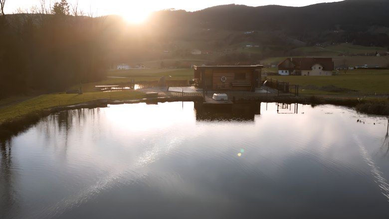 Ein Teich mit einem Holzgebäude am Ufer, umgeben von Hügeln und Bäumen im Sonnenuntergangrgang.