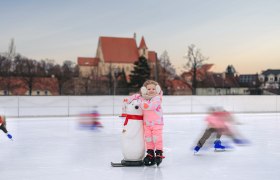 Ein Kind in pinker Kleidung steht auf einer Eisbahn mit einem Pinguin-Hilfsmittel. Im Hintergrund ist eine Kirche zu sehen.