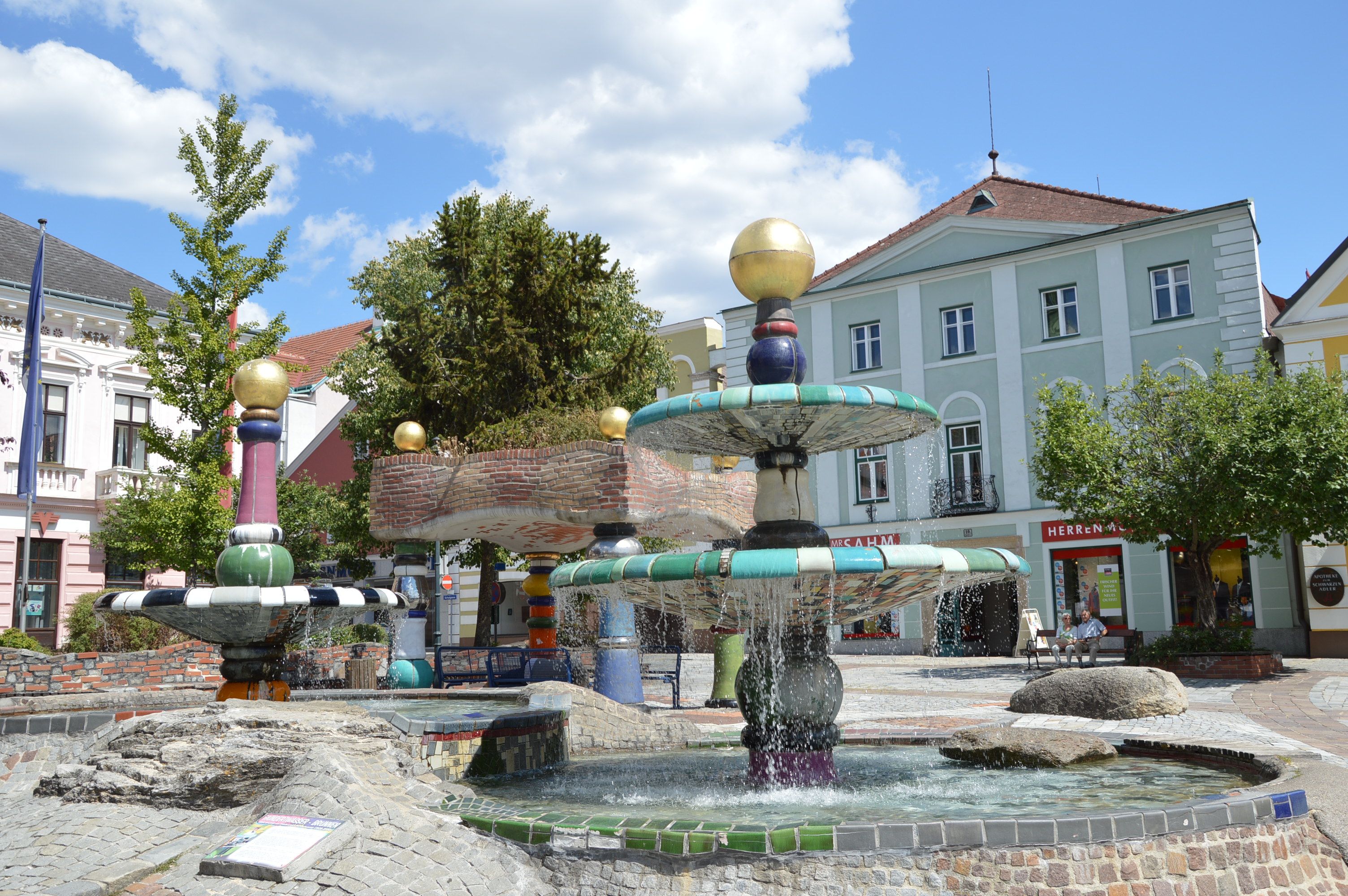 Der Hundertwasserbrunnen inmitten von der Stadtgemeinde Zwettl. Nebenbei Häuser in verschiedenen Farben.