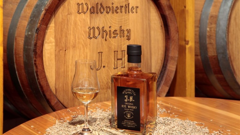 Flasche Waldviertler Whisky und Glas auf Tisch mit Getreide vor Holzfass.