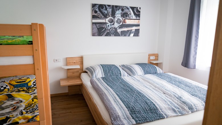 Schlafzimmer mit Doppelbett, Etagenbett und Wandbild eines Autoteils.