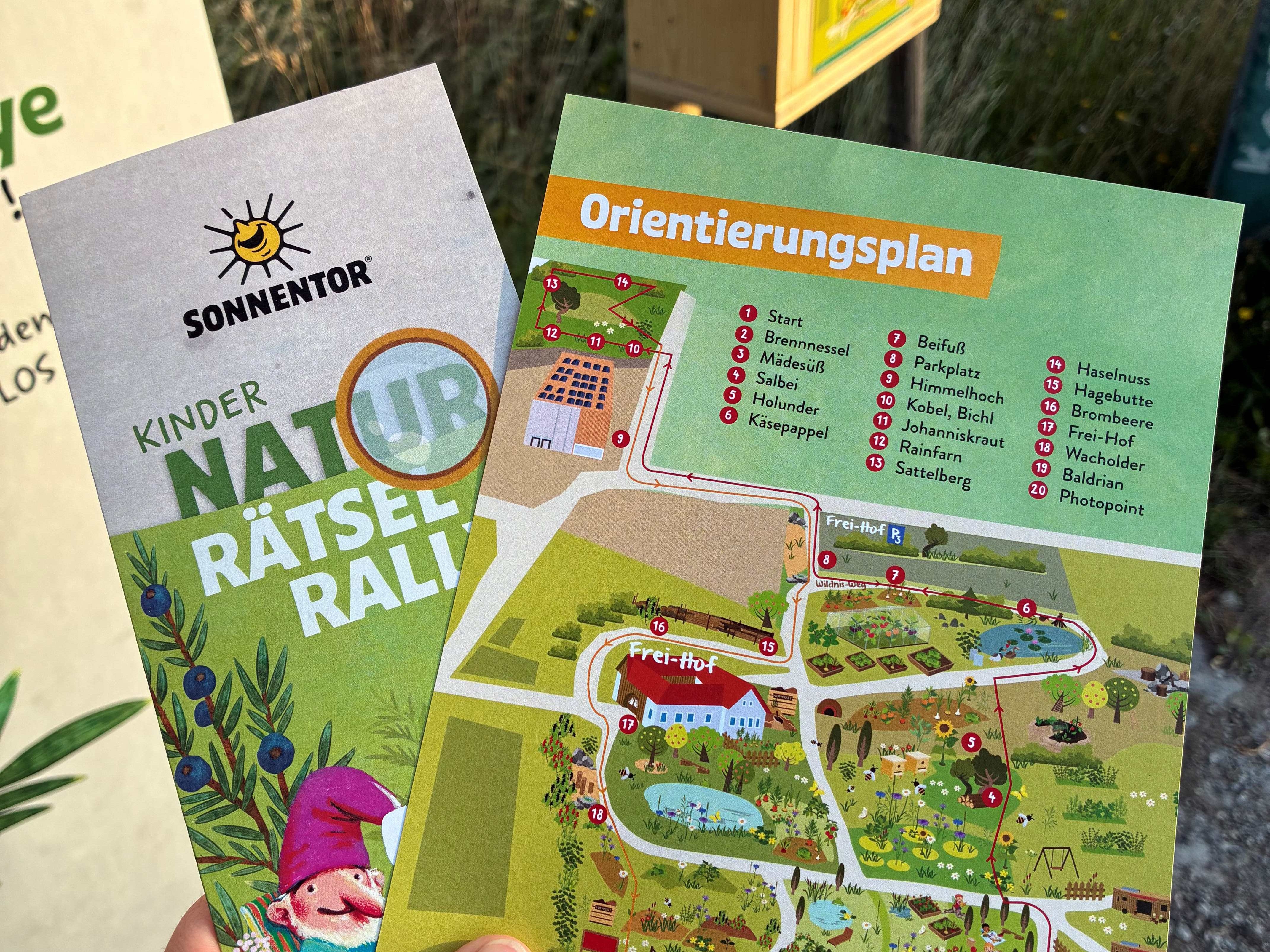 Zwei Flyer mit der Aufschrift 'Kinder Naturrätselrallye' und einem Orientierungsplan in einer Hand gehalten.
