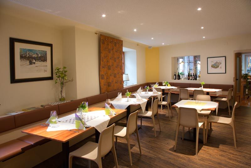 Ein moderner Speiseraum mit gedeckten Tischen, Stühlen und Dekorationen. An den Wänden hängen Bilder, und es gibt eine Fensterbank mit Weinflaschen.