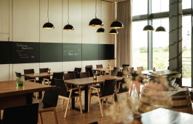 Modernes Restaurant mit Holztischen, schwarzen Stühlen und großen Fenstern. Schwarze Hängelampen und eine Tafel an der Wand.