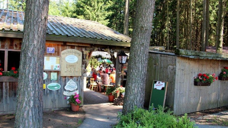 Eingang zu einer Waldschenke mit Holzhütte und Blumen.