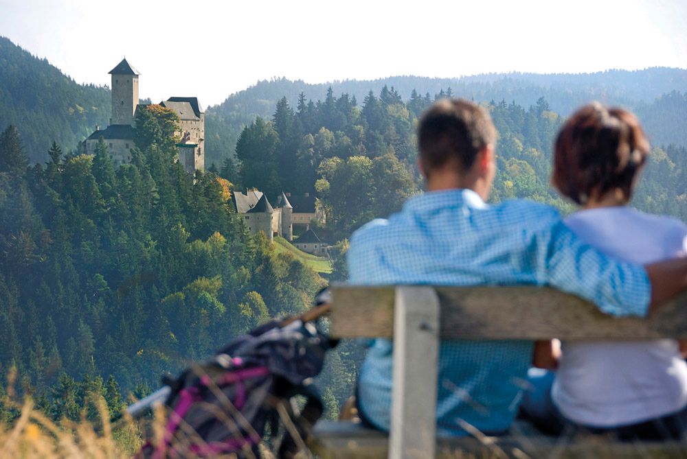 Ein Paar sitzt auf einer Bank mit Blick auf eine Burg in einer bewaldeten Landschaft.