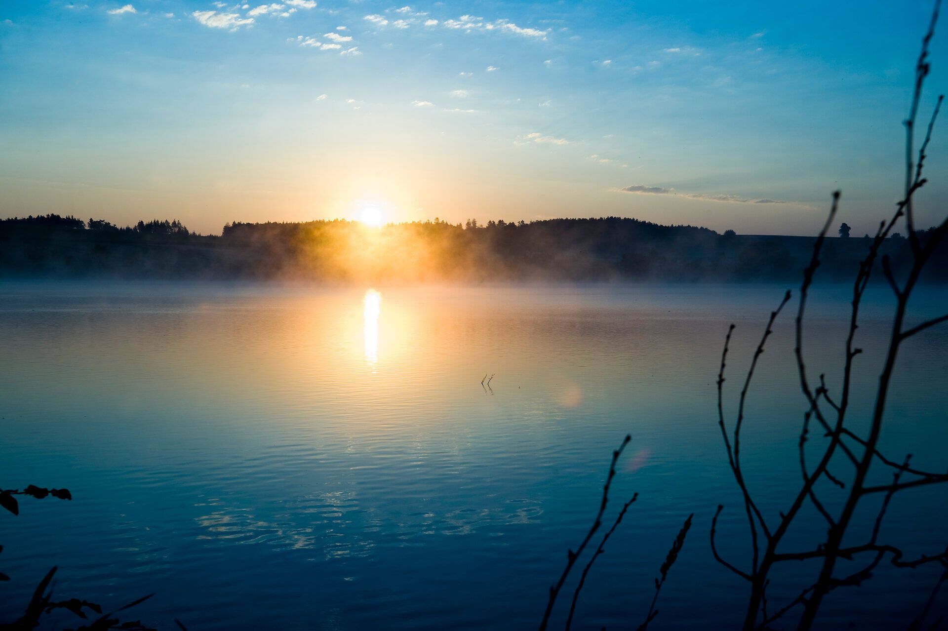 Die sanften Wellen des Sees spiegeln das goldene Licht der aufgehenden Sonne wider, während der Morgennebel sanft über die Wasseroberfläche zieht. Umgeben von üppigem Grün und dem Gesang der Vögel, lädt diese friedliche Szenerie dazu ein, die Ruhe der Natur zu genießen und den Tag mit einem Gefühl der Erneuerung zu beginnen.