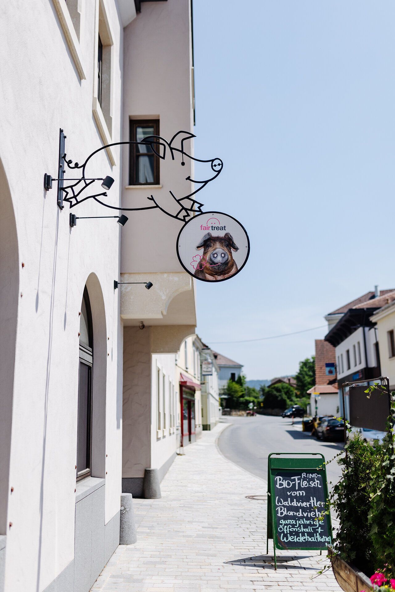 Ein einladender Ab-Hof-Laden, der mit einem charmanten, handgefertigten Schild in Form eines Schweins auf sich aufmerksam macht. Die sonnige Atmosphäre und die malerische Umgebung laden dazu ein, regionale Köstlichkeiten zu entdecken und die frische Landluft zu genießen.