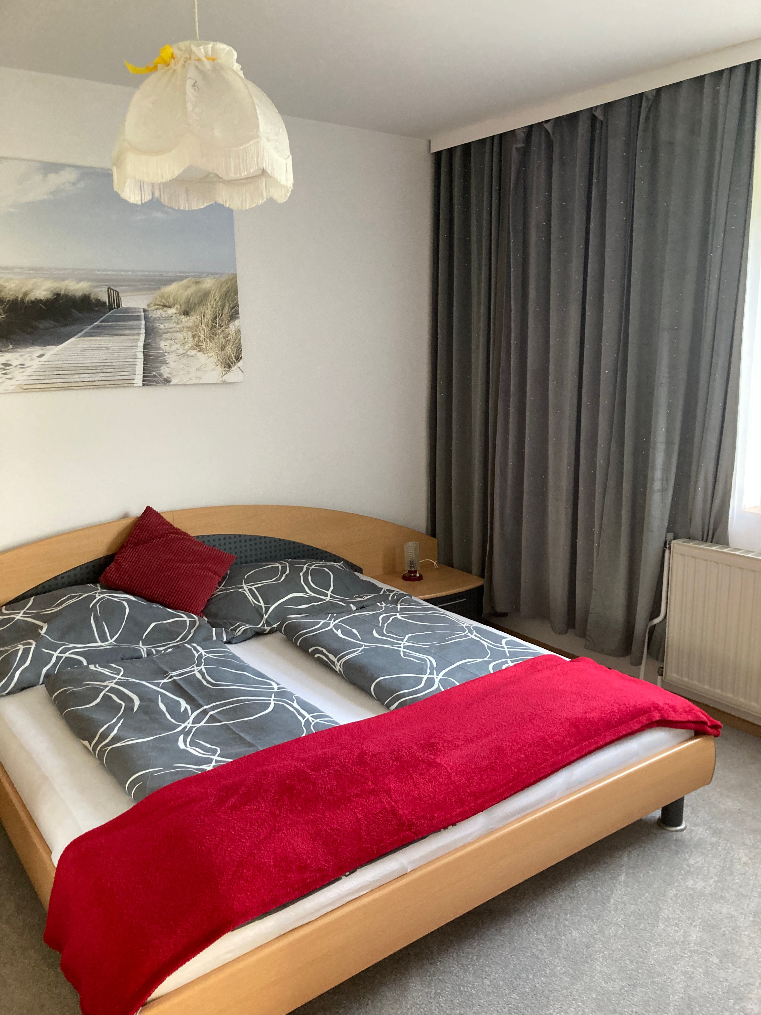 Schlafzimmer mit Doppelbett, grauer Bettwäsche, rotem Überwurf und Kissen, Lampe und Strandbild an der Wand.