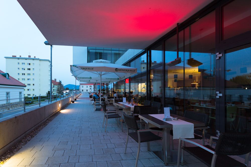 Terrasse eines Restaurants mit Tischen und Stühlen, beleuchtet von roten Lichtern, mit Blick auf Gebäude im Hintergrund.