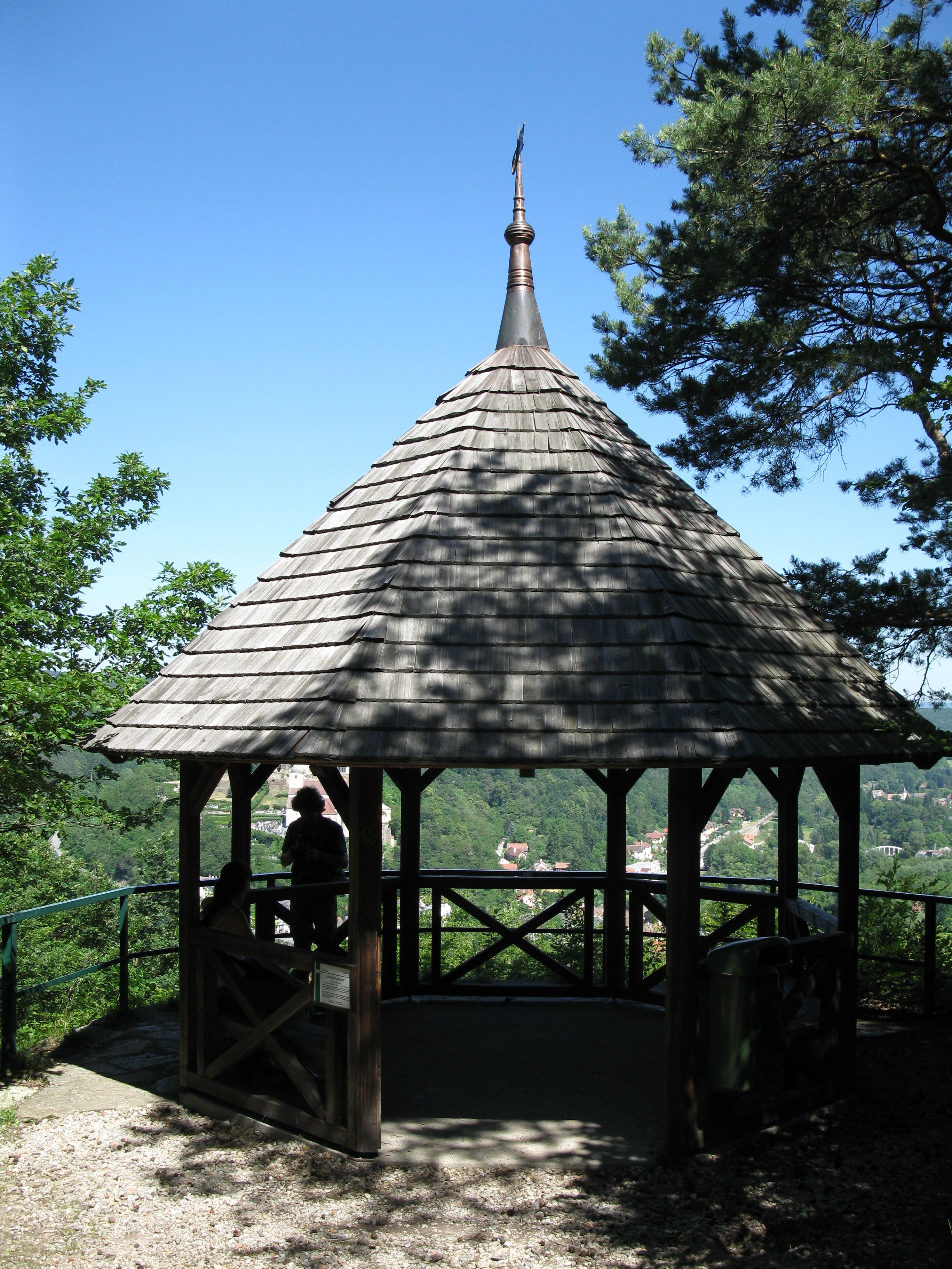 Ein hölzerner Pavillon mit spitzem Dach auf einem Hügel, umgeben von Bäumen, mit Blick auf eine grüne Landschaft.