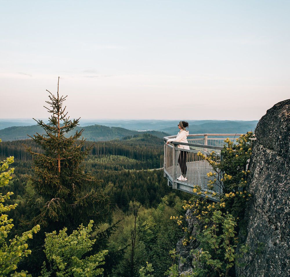 Ein atemberaubender Ausblick eröffnet sich über die sanften Hügel und dichten Wälder, während die Abendsonne den Himmel in sanften Pastelltönen färbt. Die frische Bergluft und das sanfte Rascheln der Blätter laden dazu ein, einen Moment innezuhalten und die Schönheit der Natur zu genießen.