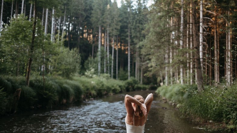 Eine Person steht in einem Fluss in einem Wald und streckt die Arme nach oben.