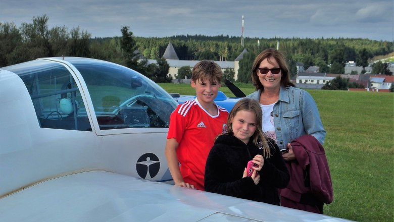 Three people are standing next to a small airplane on an airfield with a green meadow and a cloudy sky in the background.