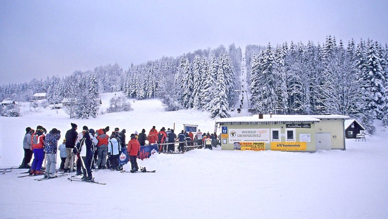Skifahrer stehen in einer Schlange vor einem Skilift in einer verschneiten Winterlandschaft.