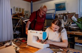 Eine Frau näht an einer Nähmaschine, während ein älterer Mann daneben steht und lächelt.