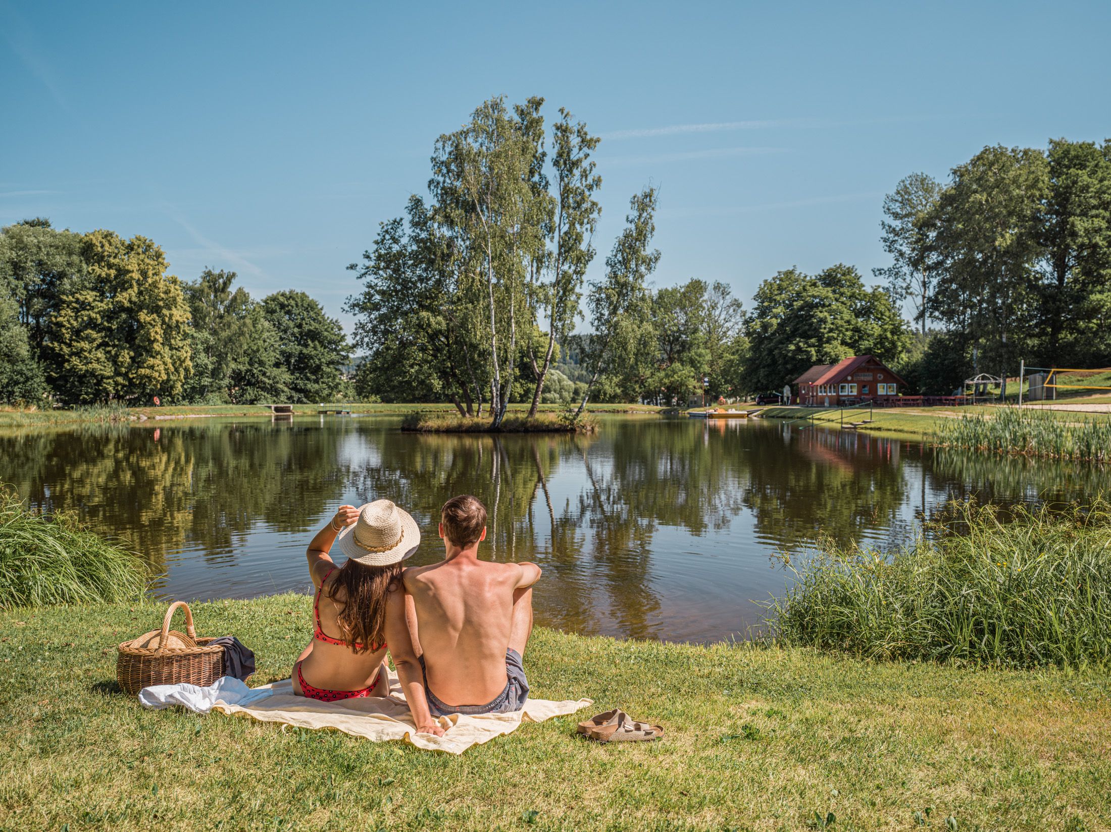 Ein Paar sitzt auf einer Decke am Ufer eines Sees, umgeben von Bäumen und einem roten Gebäude im Hintergrund.